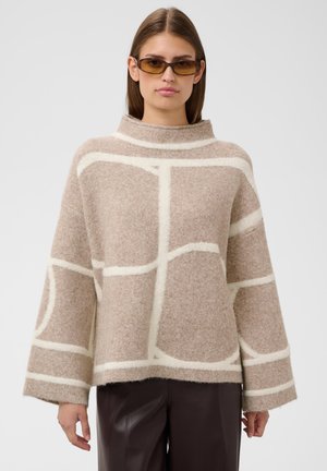 Femme portant un pull oversize beige et blanc à motifs avec de larges manches, pantalon en cuir marron et lunettes de soleil teintées ambre, debout sur fond blanc.