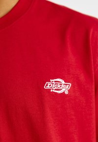 Sarkana kokvilnas T-kreklu ar baltu izšūtu "Dickies" logotipu kreisajā krūtīs, ar gludu, vieglu tekstūru.