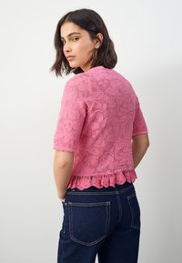 Rosa Spitzenoberteil mit floralem Muster, kurzen Ärmeln und einem gewellten Saum. Der Rücken ist etwas länger und wird mit dunklen Jeans getragen.