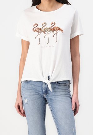 Witte top met korte mouwen en een strik aan de voorkant, met drie met pailletten versierde flamingo's in roségoud, brons en koper boven het woord "FLAMAZING."
