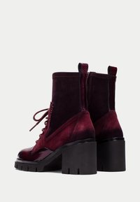 Botas de tobillo burdeos con un frontal de charol brillante y parte trasera de ante, diseño con cordones, gruesos tacones negros de bloque y texturas contrastantes.