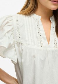 Camicia bianca in cotone con scollo a placket, delicata rifinitura in pizzo, maniche corte arricciate e pieghe sottili sul davanti.