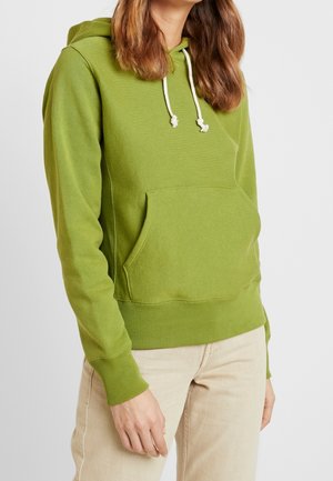 Žena v světle zelené mikině s bílými šňůrkami a béžových kalhotách stojí před jednoduchým světlým pozadím.