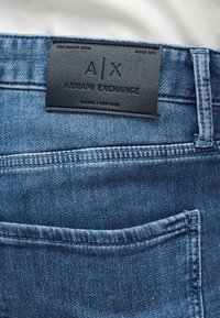 Κοντινή λήψη πίσω τσέπης μπλε τζιν με ραμμένη μαύρη δερμάτινη ετικέτα Armani Exchange πάνω από την τσέπη.