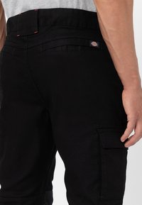 Black cargo pants with a smooth texture, featuring a side pocket, red stitching accents, and a logo patch on the back waistband.