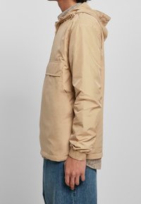 Giacca leggera beige con cappuccio, zip frontale, polsini elastici e tasca sul petto. Tessuto liscio con finitura leggermente lucida.