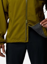 Person wearing olive green jacket and black pants, holding open jacket hem to show inner mesh lining with hexagonal pattern.
