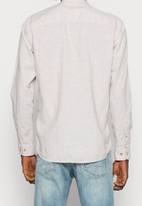 Hellgraues Hemd mit langen Ärmeln, das eine subtile Textur und goldene Knopfakzente aufweist. Getragen mit blauen Jeans.