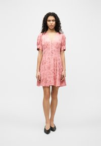 Γυναίκα με σγουρά μαλλιά που φοράει ροζ λουλουδάτο φόρεμα με κοντά μανίκια και κουμπιά μπροστά, καθώς και μαύρα μυτερά παπούτσια, όρθια.