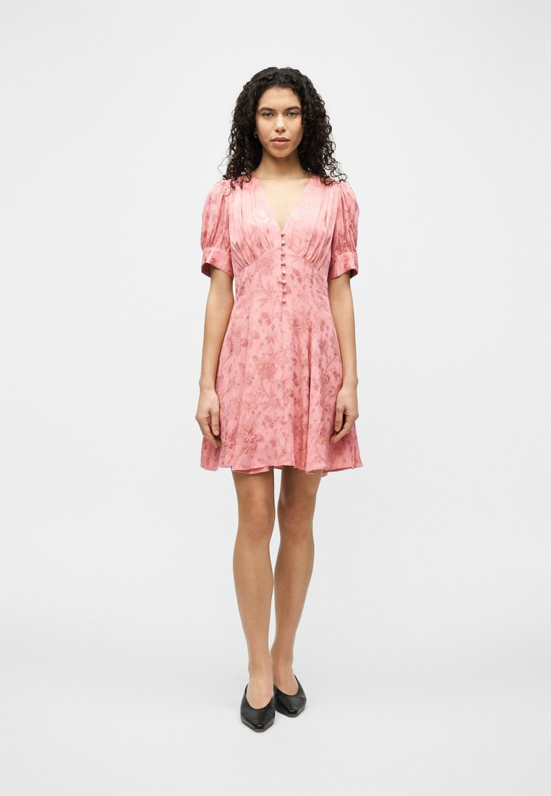 Γυναίκα με σγουρά μαλλιά που φοράει ροζ λουλουδάτο φόρεμα με κοντά μανίκια και κουμπιά μπροστά, καθώς και μαύρα μυτερά παπούτσια, όρθια.