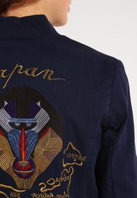 Námorně modrá bombarďák s žebrovaným límcem. Na zadní straně se nachází vícebarevná vyšívaná grafika babouna a názvy měst ve zlaté nitě.