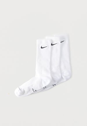 Trois paires de chaussettes blanches à tige courte avec logos Nike Swoosh noirs empilées sur une surface blanche plate.