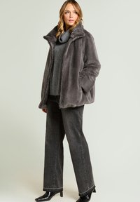 Grijze jas van nepbont met een hoge kraag, gecombineerd met een grijze trui en donkergrijze jeans met wijde pijpen. Zwarte enkellaarsjes maken de outfit compleet.