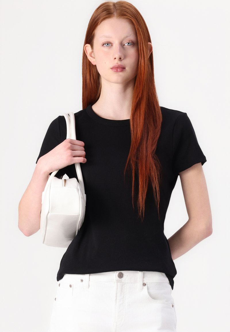 Czarny bawełniany t-shirt z krótkim rękawem, połączony z białą skórzaną torbą z zamkiem błyskawicznym i zorganizowanym fasonem. Modelka trzyma torbę.