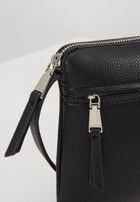 Bolso bandolera negro de cuero con un acabado texturizado, cremallera plateada y acentos de hardware metálico. Cuenta con una correa desmontable y un bolsillo frontal.