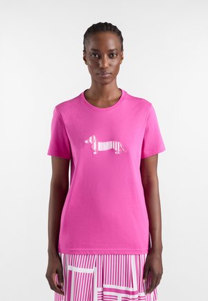 Donna che indossa una maglietta rosa brillante con una grafica di un bassotto a strisce e una gonna abbinata a righe rosa e bianche, in piedi su uno sfondo bianco.