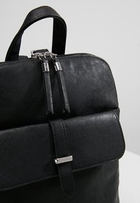 Mochila de cuero negro con asa superior, tiradores de doble cremallera, bolsillo en la parte frontal con tapa y cierre de hebilla plateada sobre un fondo claro.
