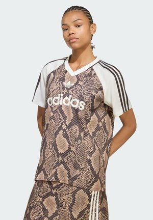 Persona con capelli intrecciati che indossa una maglietta e pantaloncini Adidas con motivo in pelle di serpente, in piedi davanti a uno sfondo chiaro e uniforme.