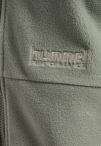 Close-up van groen fleece-stof met het geborduurde "hummel"-logo boven een ritssluiting.