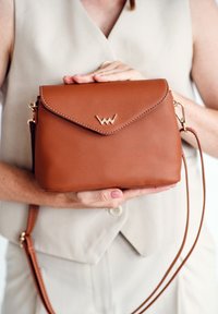 Braune Lederhandtasche mit einem goldenen Emblem, strukturierter Form, Klappenoberseite und einem abnehmbaren Schulterriemen. Texturierte Oberfläche mit Nähdetails.