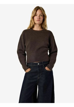 Femme aux cheveux blonds portant un pull à manches longues marron foncé et un jean large foncé, debout avec les mains dans les poches sur fond blanc.