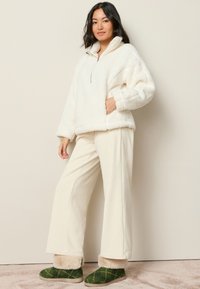 Hvid fleecepullover med halvlynlås, kombineret med cremede viddebenbukser og grønne ternede hjemmesko med pelskant.