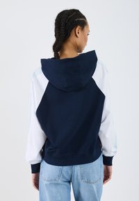 Felpa con cappuccio blu navy con accenti bianchi e azzurri chiari. Presenta una vestibilità rilassata, polsini a coste e una tasca a marsupio.