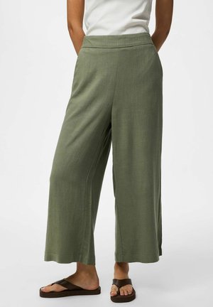 Personne portant un pantalon large court vert olive, un haut blanc sans manches et des sandales marron à brides, debout devant un fond blanc uni.