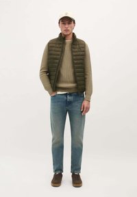 Vesta matlasată verde peste un pulover tricotat bej, asortată cu blugi de culoare albastru deschis și adidași maro. O șapcă de culoare deschisă finalizează ținuta.