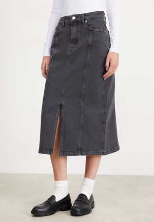 Frau trägt einen schwarzen Denim-Midirock mit vorderem Schlitz, weiße Socken, schwarze Loafers und ein weißes Langarmshirt vor einem einfarbigen Hintergrund.