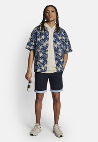 Tenue décontractée comprenant une chemise à manches courtes bleu floral, un hoodie beige, un short navy avec des poignets bleu clair et des baskets beiges. Lunettes de soleil à la main.