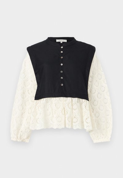 Veste noire sans manches raccourcie portée sur une blouse blanche à manches longues en dentelle avec boutons sur le devant et poignets froncés, sur un fond neutre.