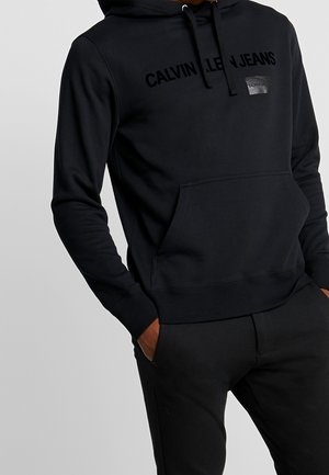 Hombre con sudadera negra de Calvin Klein Jeans con bolsillo delantero y capucha con cordón, manos en los bolsillos del pantalón, visible desde el cuello hasta las caderas.