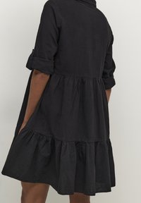 Schwarzes Baumwollkleid mit einem geknöpften Kragen, langen Ärmeln, die an den Bündchen hochgekrempelt sind, und einem abgestuften, ausgestellten Rock. Strukturierter Stoff mit Taschen.