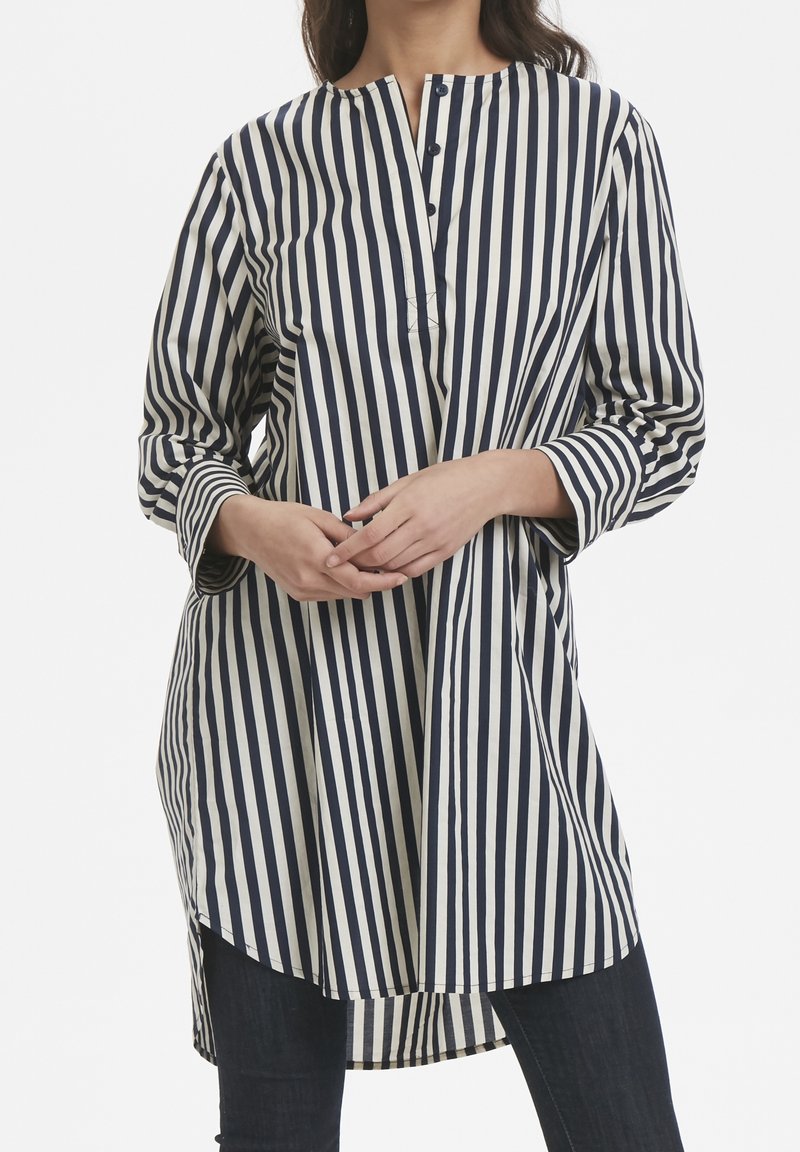 Túnica de rayas azul marino y crema con cuello redondo, tapeta de botones y mangas largas, con dobladillo trasero más largo y textura de tela suave.