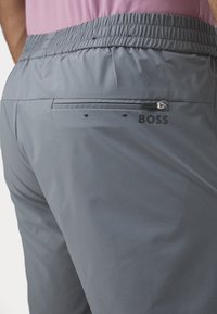Pantalones cortos deportivos grises con cintura elástica, que cuentan con un bolsillo con cremallera y un detalle de logo en la parte posterior. Textura de tela suave.