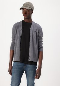 Cardigan gri cu nasturi și un mic logo, purtat peste un tricou negru, asortat cu blugi blue și o șapcă gri cu accente uzate.