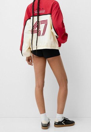 Une personne aux longues tresses porte une veste rouge et crème avec le numéro « 47 » dans le dos, un short noir, des chaussettes blanches et des baskets noires à semelles en gomme.