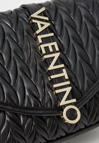 Borsa in pelle trapuntata nera con logo "VALENTINO" dorato su una tracolla decorativa. Texture liscia con dettagli in rilievo e motivi.