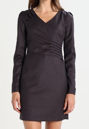 Frau trägt ein eng anliegendes, dunkel gefärbtes Langarmkleid mit dezentem geometrischem Muster und V-Ausschnitt, steht vor einem schlichten Hintergrund.