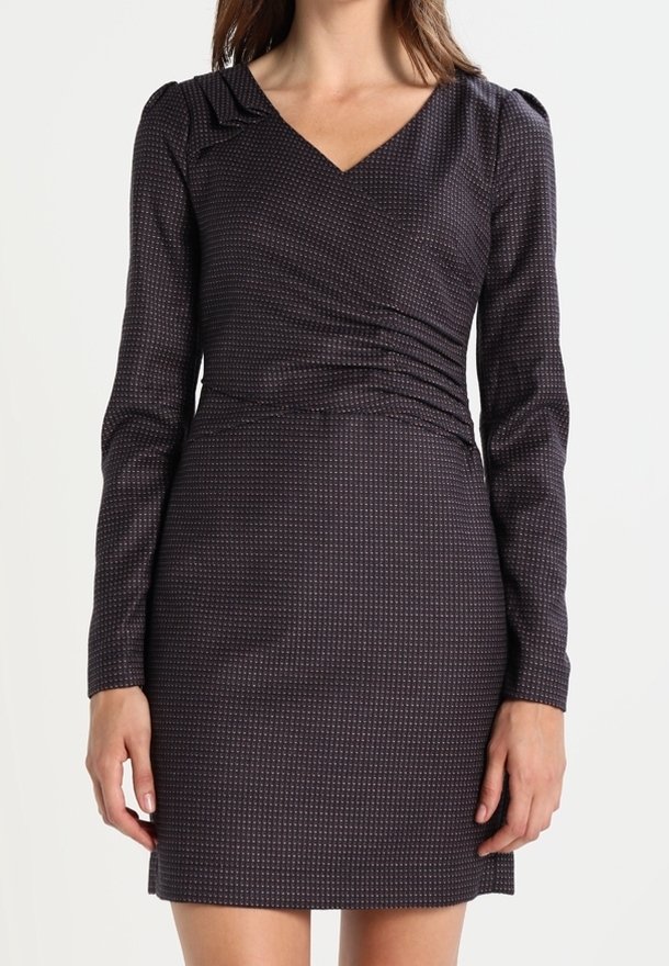 Femme vêtue d'une robe ajustée sombre à manches longues avec un motif géométrique subtil et un col en V, debout devant un fond uni.