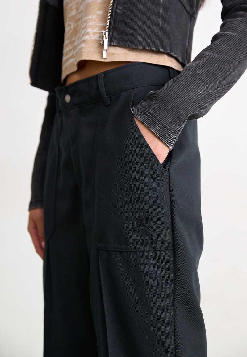 Schwarze weit geschnittene Hose aus robustem Stoff, mit Seitentaschen und einem dezenten schwarzen Jordan-Logo, das links eingestickt ist.