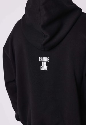 Felpa nera con cappuccio realizzata in materiale morbido, caratterizzata da un grande cappuccio e da una scritta bianca "CHANGE THE GAME" sul retro. Design semplice, vestibilità casual.