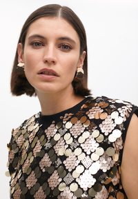 Black top adorned with overlapping gold, rose gold, and silver sequins in various sizes. Model wears large, geometric earrings.