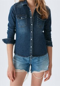 Kvinna som bär en mörk denimskjorta med knappar och uppvikt ärm, samt ljusblå slitna denimshorts mot en enfärgad bakgrund.