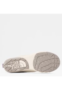 Zapatilla blanca con una suela de goma beige texturizada. Presenta el logo "The North Face" en blanco en la parte inferior y un acento morado en la parte trasera.