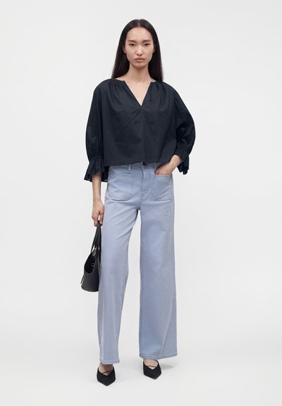 Femme portant un chemisier noir ample, un jean bleu clair à jambes larges, des chaussures noires pointues, tenant un sac à main noir avec une main dans la poche.