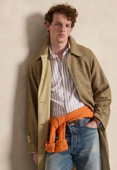 Junger Mann mit lockigem Haar, der einen karierten Mantel, ein gestreiftes Hemd, einen orangefarbenen Pullover, der an der Taille gebunden ist, blaue Jeans und eine Armbanduhr trägt, steht an einer Wand.