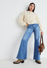 Cardigan lavorato a maglia crema con motivo testurizzato, jeans azzurri a zampa d'elefante e stivali beige con tacco. La borsa a spalla marrone crea un contrasto.