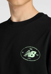 T-shirt noir à manches courtes avec un col rond, présentant un logo ovale vert avec l'inscription "New Balance Athletics Inc." et d'autres détails.